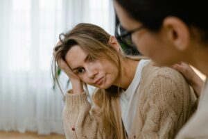 A person with long blonde hair sits in a softly lit room, comforted by another’s reassuring touch on their shoulder, reflecting support and emotional well-being.